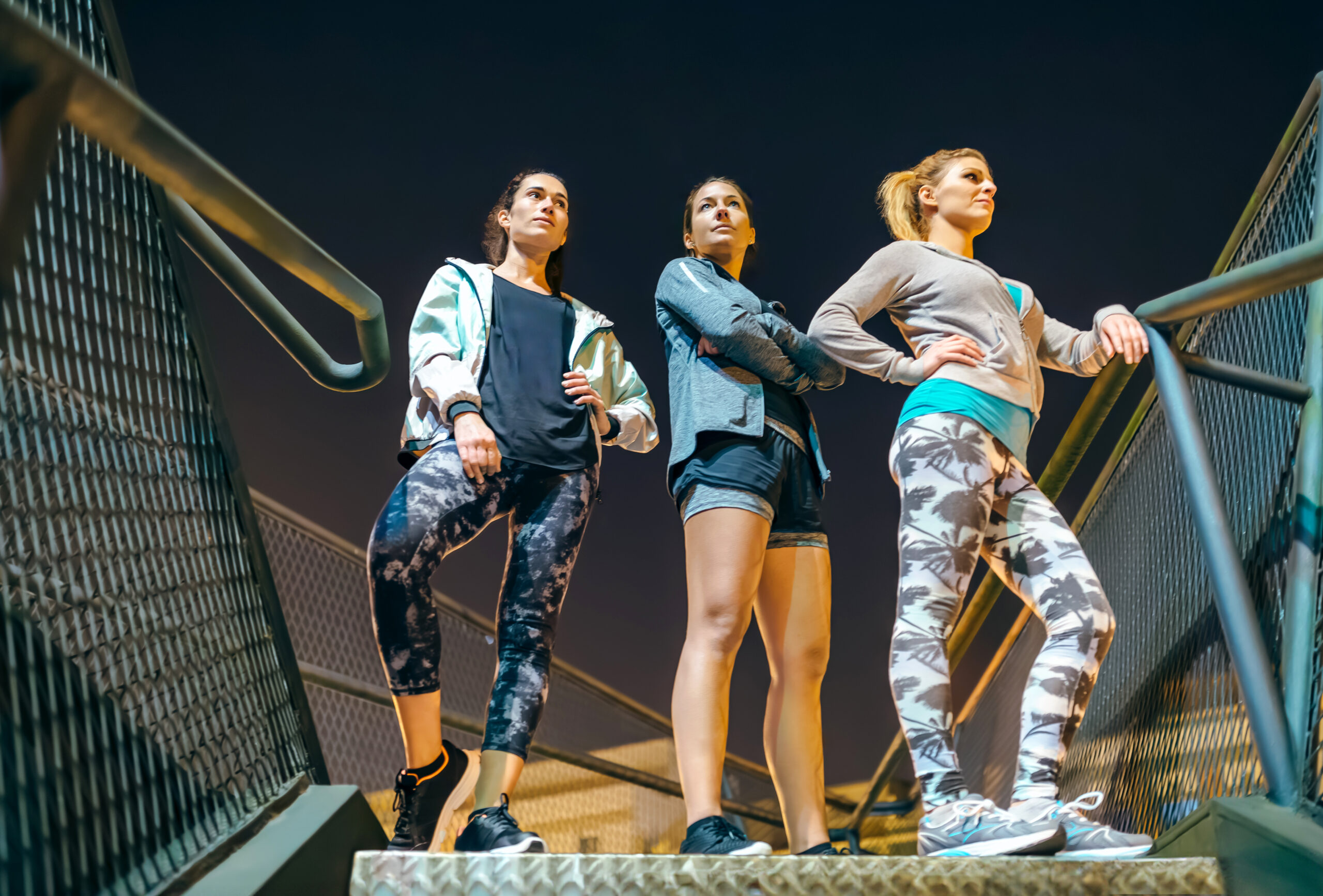 Three athletic women standing on urban stairs at night ready for an evening exercise session in the city. Group of female friends runners dressed with sportive wear over metallic staircase outdoors.