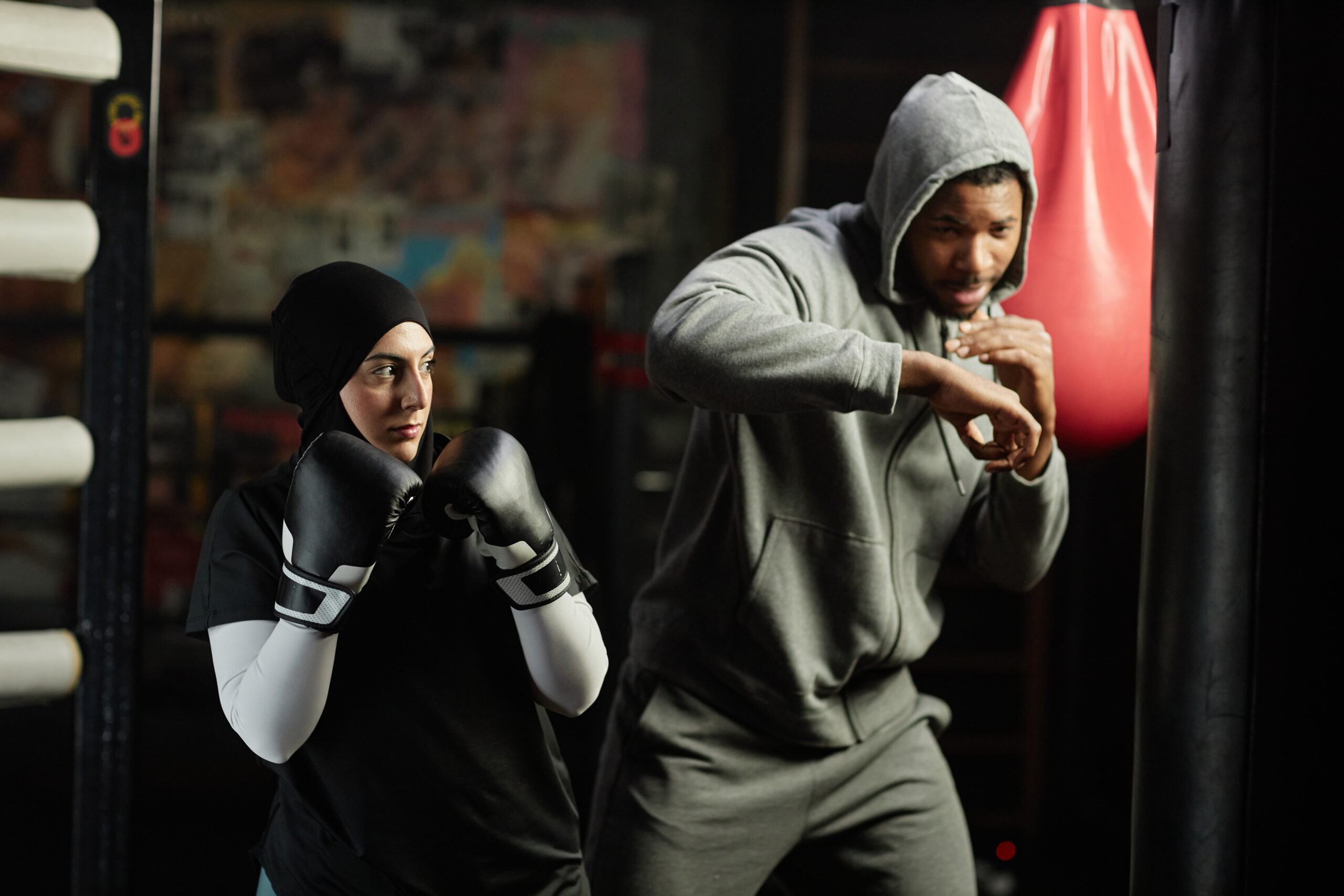 Young serioua Muslim sportswoman in boxing gloves crossing in front of herself and looking at punching bag before hitting it during training